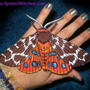 May include: A brown and white moth with orange and blue accents. The moth is embroidered and has a 3D effect. The moth is on a hand.
