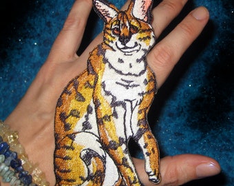 May include: An embroidered patch of a brown and white tabby cat sitting with its tail curled around its legs. The cat has a white chest and a black nose.