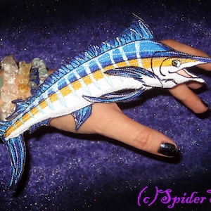 Può includere: Una toppa ricamata a forma di pesce marlin blu e bianco con accenti gialli. La toppa è su una mano con un braccialetto di pietre preziose verdi e trasparenti.