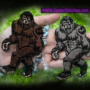 May include: Two embroidered iron-on patches of Bigfoot, one brown and one grey, with a hairy face and a walking pose. The patches are on a hand with a bracelet.