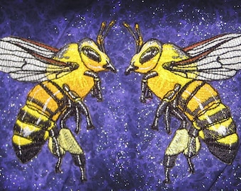 May include: Two embroidered bees facing each other on a purple background with a glittery texture. The bees are yellow and black with detailed wings.