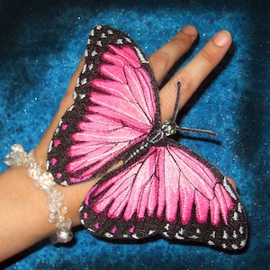Puede incluir: Una mariposa rosa y negra con un diseño bordado detallado. La mariposa está en una mano con una pulsera de cristal transparente.