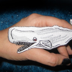 Puede incluir: Un parche blanco bordado de una ballena de esperma con un ojo rojo, en la mano de una persona.