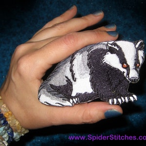 May include: A black and white embroidered patch of a badger with orange eyes. The patch is being held in a hand with blue nail polish.