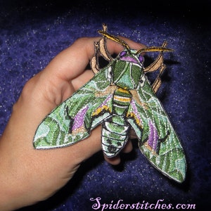 Könnte beinhalten: Grüne, violette und goldene gestickte Motten-Aufnäher mit detailliertem Design. Die Motte hat große Flügel und einen langen Körper. Der Aufnäher ist perfekt, um Kleidung, Taschen oder anderen Accessoires eine einzigartige Note zu verleihen.