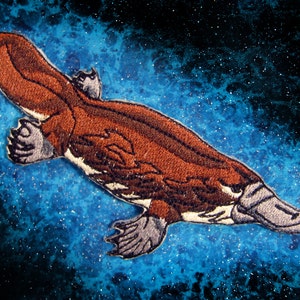 May include: A brown and gray embroidered patch of a platypus swimming. The platypus is on a blue background with white speckles.