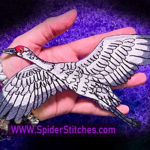 May include: A white and gray embroidered iron-on patch of a crane in flight. The crane has a red head and a black outline. The patch is approximately 5 inches wide and 7 inches long. The patch is on a hand with a purple background. The text "WWW.SpiderStitches.com" is visible in the bottom of the image.