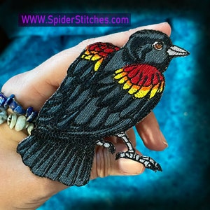 Op de afbeelding: Gestikt patch van een zwarte vogel met rode en gele accenten op zijn vleugels. De vogel zit op een hand, met de tekst "WWW.SpiderStitches.com" zichtbaar op de achtergrond.