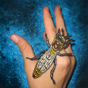 May include: A yellow and black embroidered insect patch with white wings. The patch is on a hand.