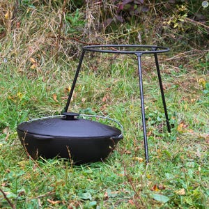 May include: A black cast iron pot with a lid sits on a black metal tripod stand. The pot and stand are on a bed of green grass.