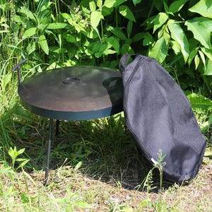 May include: A black metal fire pit with a circular cooking surface and three legs. A black carrying bag rests beside the fire pit. The fire pit is outdoors, surrounded by green grass and foliage.