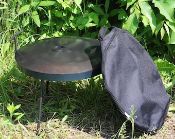 Peut inclure: Un brasero en métal noir avec une surface de cuisson circulaire et trois pieds. Un sac de transport noir est posé à côté du brasero. Le brasero est à l'extérieur, entouré d'herbe verte et de feuillage.