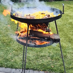 Puede incluir: Un brasero de metal negro con una rejilla de cocción redonda y una rejilla inferior para el fuego. El brasero está instalado en una superficie cubierta de hierba y tiene fuego ardiendo en su interior. Hay patatas y otros alimentos cocinándose en la rejilla superior.