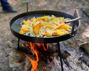 Peut inclure: Une poêle de cuisson ronde en métal noir avec des poignées, soutenue par un trépied, cuisinant des pommes de terre et des oignons au-dessus d'un feu de camp. Les pommes de terre sont dorées et les oignons sont blancs translucides.