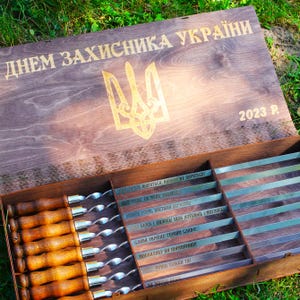 Puede incluir: Una caja de madera con una inscripción dorada que dice "Dnem Zahisnika Ukraini" (Día del Defensor de Ucrania) y el año 2023. La caja contiene un juego de 10 pinchos de metal con mangos de madera. Los pinchos están grabados con frases ucranianas.
