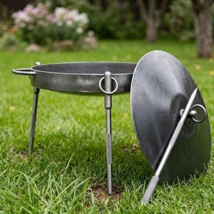 Op de afbeelding: Een donkergrijze metalen vuurplaats met bijpassend deksel, op drie poten. De vuurplaats heeft een ronde kom met een handvat en een ring aan de zijkant. Het deksel heeft een handvat en een punt.