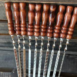 May include: A set of ten stainless steel skewers with twisted shafts and varnished brown wooden handles. The handles have a bulbous shape and are arranged in a row, with the skewers pointing downwards. The skewers are designed for grilling.