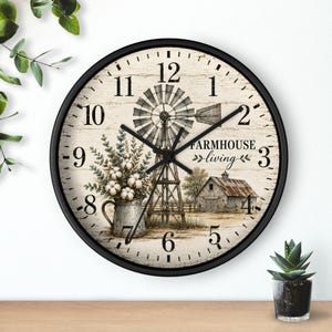 May include: A round wall clock with a black frame and a cream-colored face featuring a printed scene of a windmill, a barn, and a pitcher of cotton stems. The text "FARMHOUSE living" is printed in black, and the clock has black hour and minute hands.