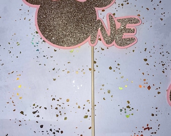 May include: A pink and gold glitter Minnie Mouse cake topper with the word "one" on the side. The topper is attached to a wooden stick.