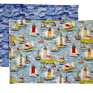 May include: Two rectangular placemats. One features a blue ocean wave pattern. The other has a light blue background with sailboats, lighthouses, and small houses. The lighthouses are red, white, and yellow.