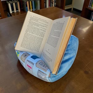 May include: A soft, blue, wedge-shaped book pillow featuring a printed pattern of various book covers on a grid background. An open hardcover book with yellowed pages is resting in the center groove of the pillow, held open for reading.