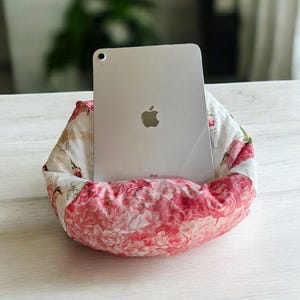 May include: A silver iPad stands in a floral fabric holder. The holder is a mix of white fabric with small floral patterns and a pink fabric with a large rose pattern. The iPad has the Apple logo on the back.
