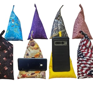 May include: Assortment of fabric phone stands in various patterns, including floral, seashell, camouflage, and an American flag design. Each stand is a triangular shape with a loop at the top, designed to hold a phone or tablet.