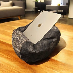 May include: A silver iPad rests in a dark gray fabric stand with a leaf pattern. The stand is a soft, cushioned design, providing support for the tablet. The stand has a camouflage pattern on one side.