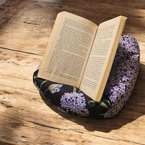 May include: An open book rests on a floral patterned book pillow. The pillow features a black background with purple and white flower designs. The book's pages are yellowed, suggesting age.