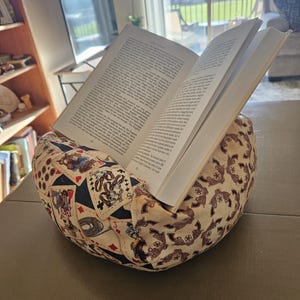 May include: A fabric book holder with a book open to page 222. The holder is patterned with playing cards, revolvers, and other western-themed designs. The book is propped up on a table.