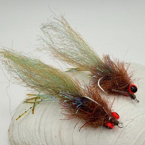 May include: Two fishing flies featuring brown synthetic fibers, long iridescent green and tan tails, and prominent red eyes with black pupils. Each fly is constructed with a metal hook and includes thin, speckled rubber legs extending outward.