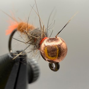 May include: Close-up of a handmade fishing fly. The fly features a copper-colored, spherical head with a yellow accent, a red band, and a body of brown and orange fibers. The hook is black.