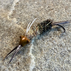 May include: Close-up of a handmade fishing fly. The fly features a dark hook, a brown head, and a body of mixed-color fibers. Two white feathers and two dark, pointed extensions are visible. The fly is designed to mimic an insect.