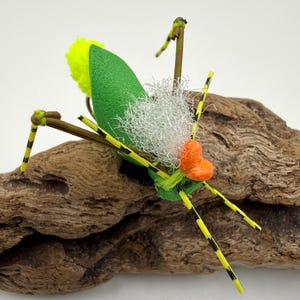 May include: A handmade fishing fly designed to resemble an insect. The fly features a green foam body, yellow and black striped legs, a yellow pom-pom, and a white fluffy material. An orange accent is also included.