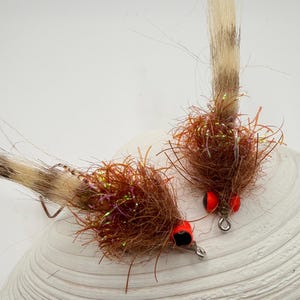 May include: Two fishing flies featuring brown, fuzzy bodies with iridescent fibers, long tan and brown striped hair tails, and prominent red eyes with black pupils. Each fly includes a small silver metal hook loop at the front for attachment.