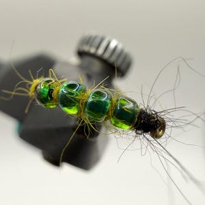 May include: A close-up of a fishing fly with four green, iridescent beads strung together. The fly is tied with yellow thread and has black and brown accents. The fly is attached to a black tool.