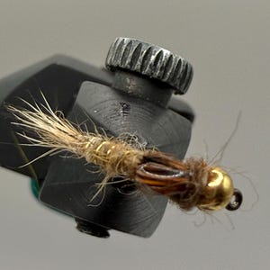 May include: Close-up of a fly fishing lure. The lure features a gold-colored bead head, brown and tan body, and a cluster of light-colored fibers. The lure is held in a black vise with a textured silver knob.
