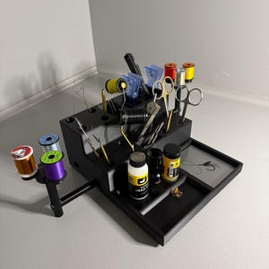 May include: Black fly tying station with a drawer, a spool holder, and various tools and materials, including thread, scissors, and bottles of fly tying head cement.