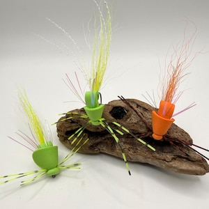 May include: Three colorful fishing flies with green, yellow, and orange accents are displayed on a piece of driftwood. Each fly features a cone-shaped body, long, thin legs, and feathery, multi-colored antennae. The flies are designed for angling.