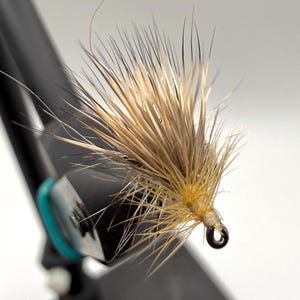 May include: Close-up of a handmade fishing fly. The fly features a cluster of tan and brown fibers, with a yellow-orange base and a small, dark hook. The fly is attached to a black tool with a teal band.