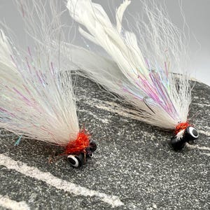 May include: Two white fishing flies with iridescent accents and red thread details. The flies have black eyes and are designed for catching fish. The flies are set against a textured gray background.