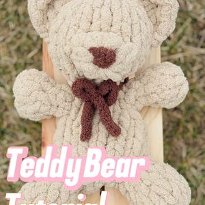 May include: A beige crochet teddy bear made of thick, soft yarn with a dark brown nose and a dark brown bow tied around its neck. The text "Teddy Bear Tutorial" is overlaid in large pink letters, with "Video & PDF" in white at the bottom.