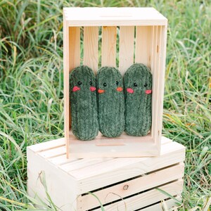 May include: Three green plush pickle toys with embroidered faces are displayed in a wooden crate. The crate is sitting on top of another wooden crate.