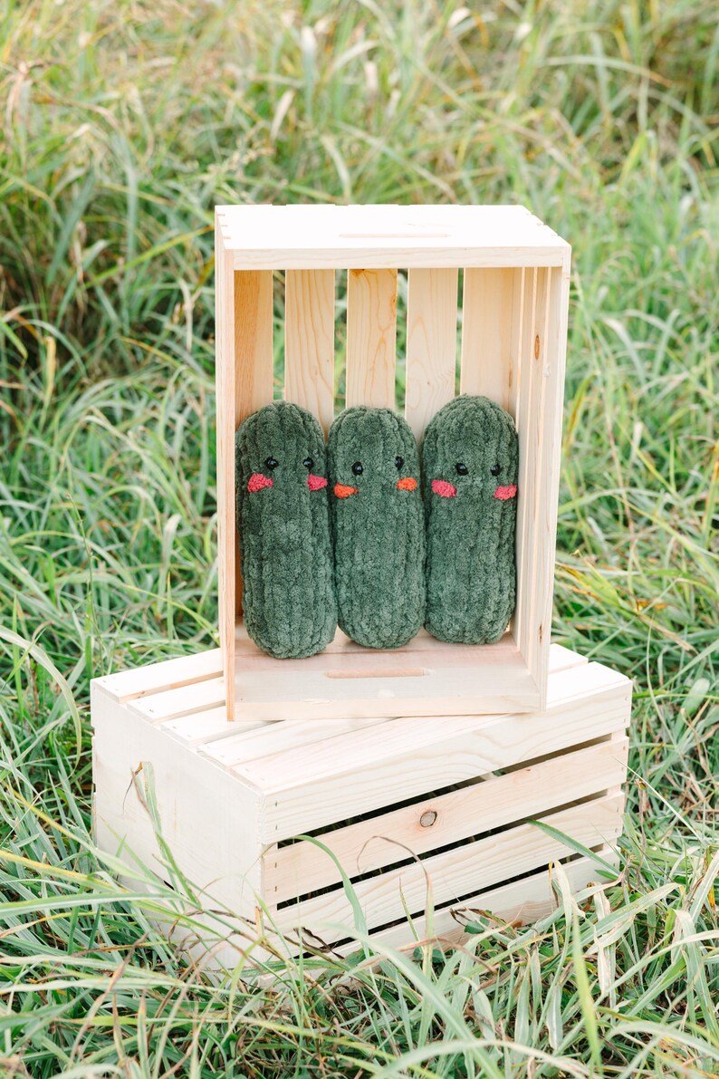 May include: Three green plush pickle toys with embroidered faces are displayed in a wooden crate. The crate is sitting on top of another wooden crate.