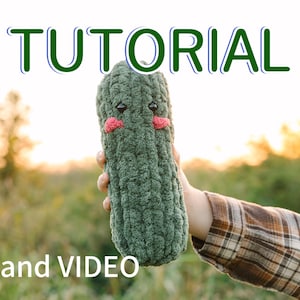 May include: A green crocheted amigurumi pickle toy with a cute face, held in a hand against a blurred background of green foliage and a sunset.
