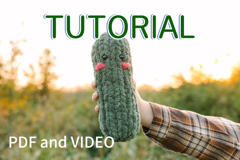 May include: A green crocheted amigurumi pickle toy with a cute face, holding in a hand against a blurred background of green foliage and a sunset.
