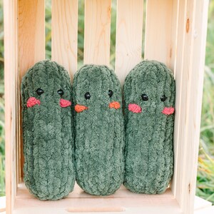 May include: Three green plush cucumber-shaped toys with embroidered eyes and pink cheeks are arranged in a wooden crate.