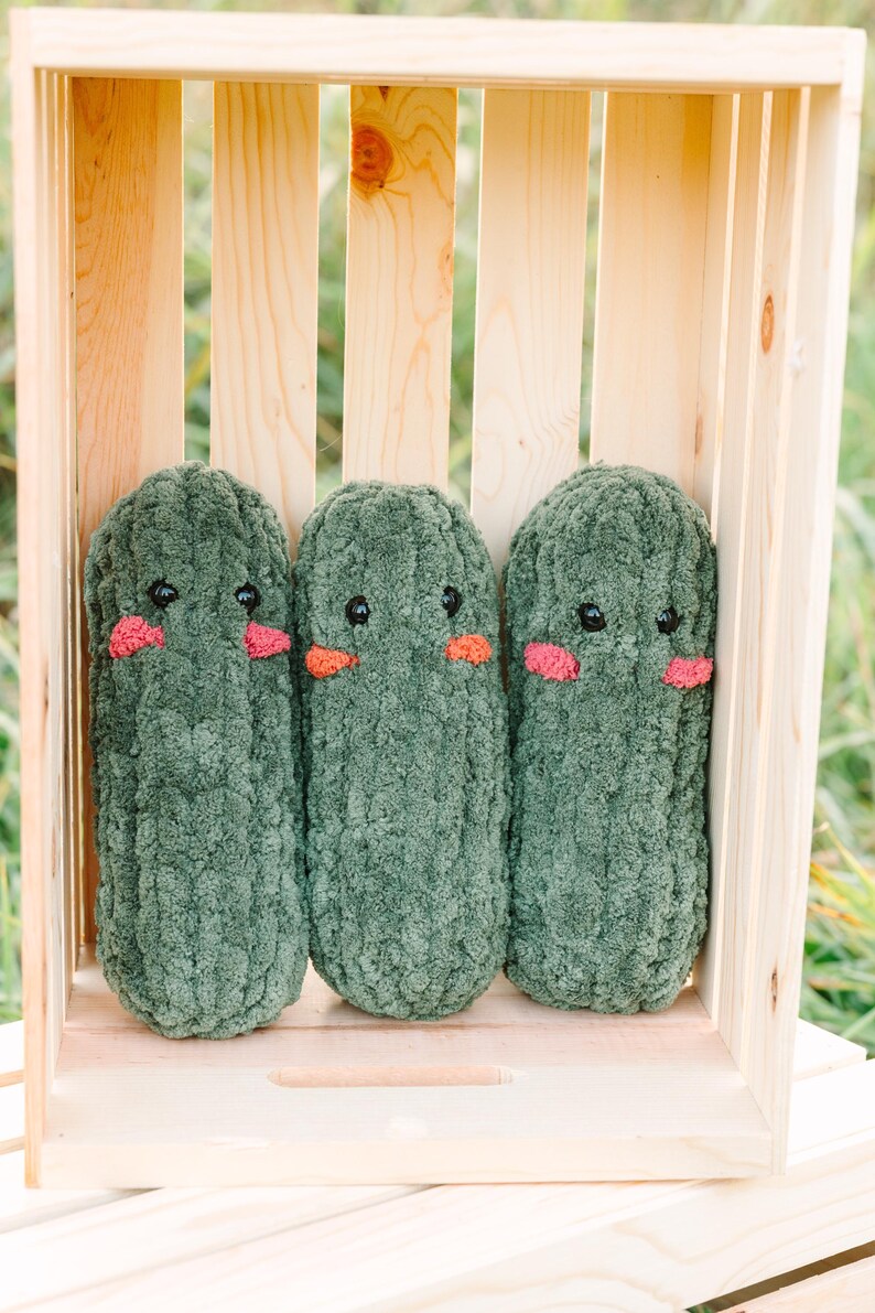 May include: Three green plush cucumber-shaped toys with embroidered eyes and pink cheeks are arranged in a wooden crate.