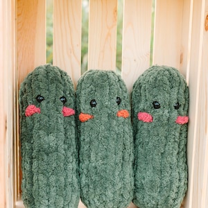 May include: Three green plush toys shaped like cucumbers with embroidered eyes and pink cheeks are arranged in a wooden crate.
