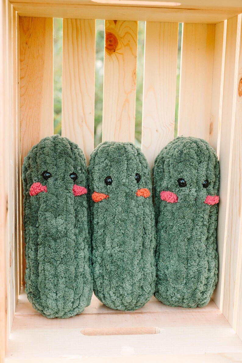 May include: Three green plush toys shaped like cucumbers with embroidered eyes and pink cheeks are arranged in a wooden crate.